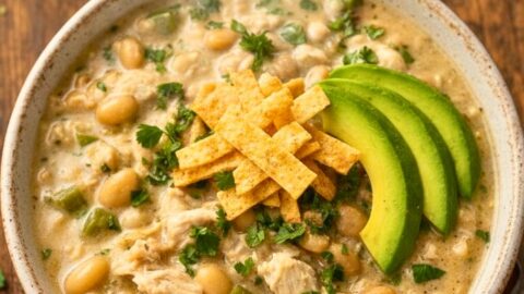 A creamy bowl of white chicken chili with tender chicken, white beans, green chilies, garnished with cilantro, avocado slices, and tortilla strips on a wooden table.